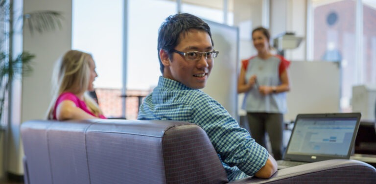 Photo of HSU Student with computer looking back at camera, while two unfocused women share conversation in the background.