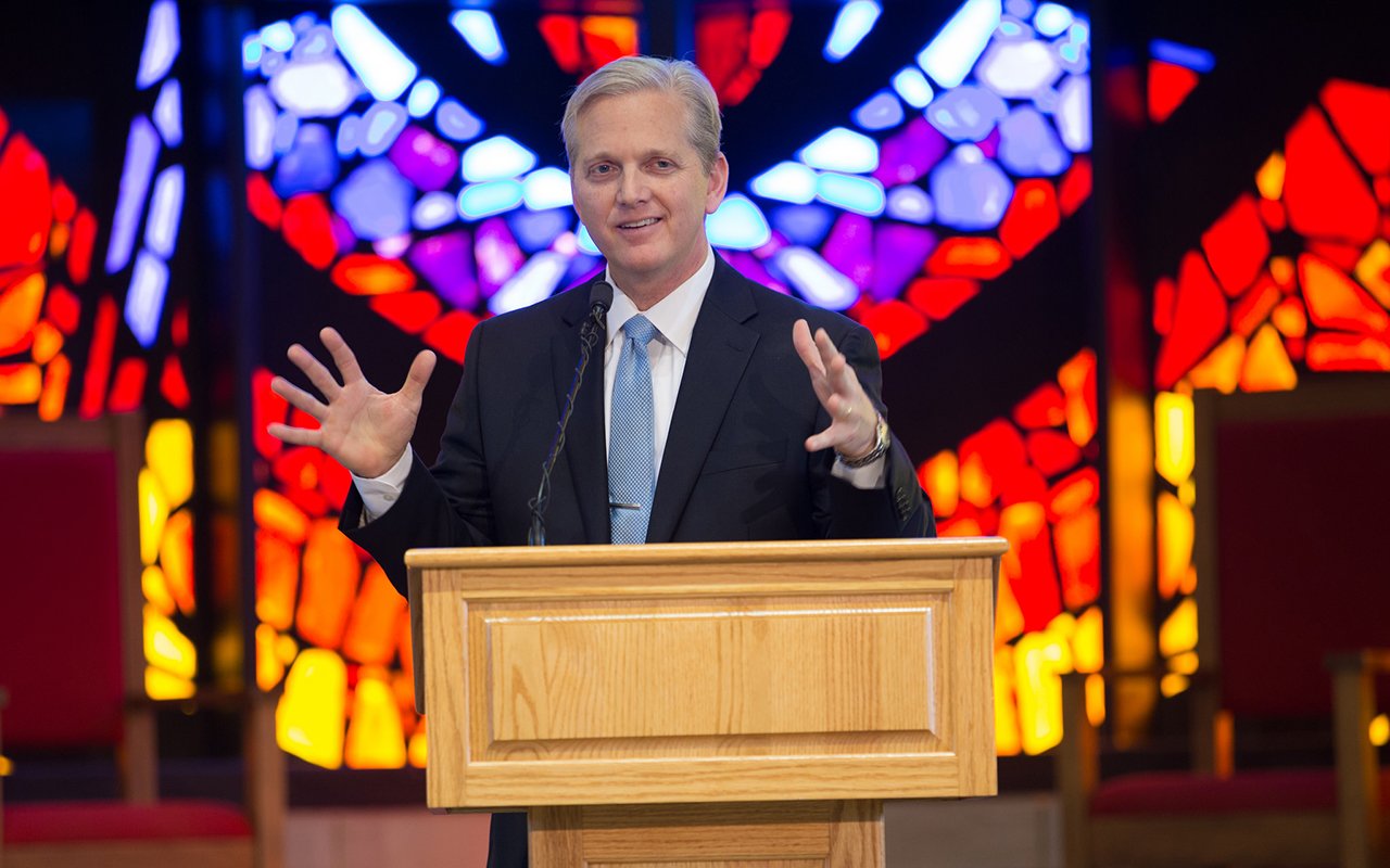 Photo of HSU President Eric Bruntmyer speaking at podium in front of Logsdon Chapel's stained glass window.