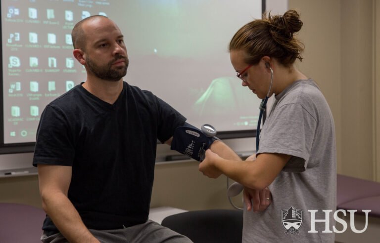 Photo of HSU student holding stethoscope end to man's inner elbow and observing her wrist watch as she takes his blood pressure.