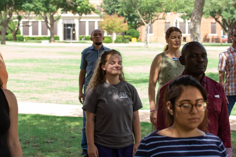Photo of Students and CEDAR participants standing and closing their eyes outdoors as they practice group yoga.