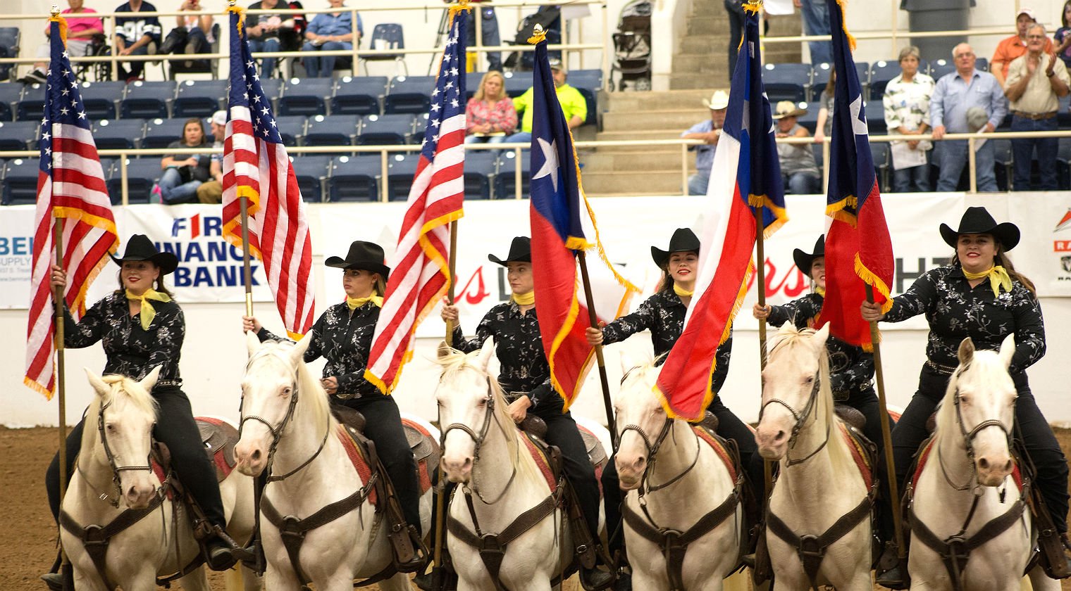 Six white horses and riders are lined up with U.S. and Texas flags.