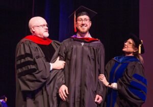 HSU graduate student at graduation standing between two professors smiling.
