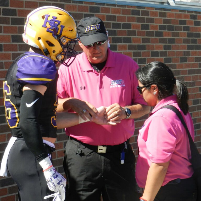 An athletic training professor wraps a football player's wrist as a student trainer observes.