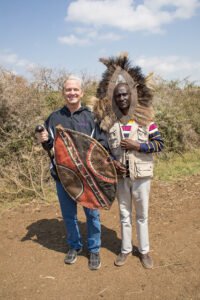 HSU President Eric Bruntmyer smiles with a Kenyan man in a headdress while holding a traditional Maasai shield.