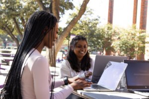 Two students sit outdoors at a campus table working on laptops together, smiling and collaborating under large trees on a sunny day.