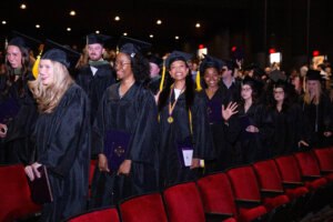 Students in graduation caps and gowns walk through an auditorium during commencement ceremony, smiling and carrying diploma covers while family and friends watch from the audience.