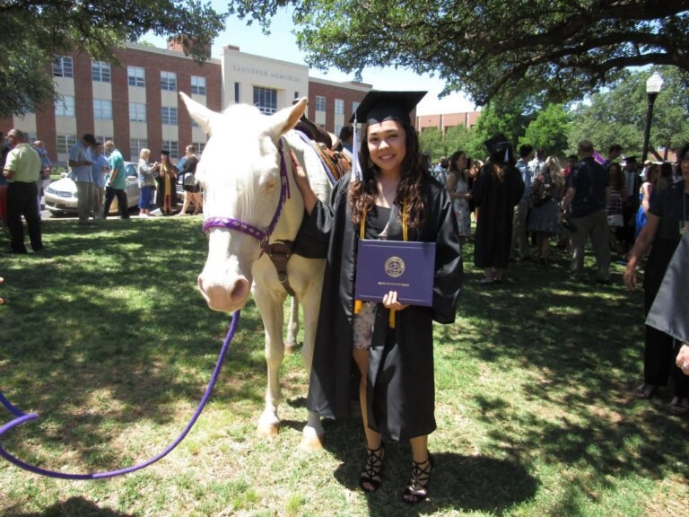 Student smiling with cap and gown on a lawn with a horse.